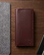 Brown leather wallet with a brand logo on a wooden surface, next to a gray felt item and a black phone.